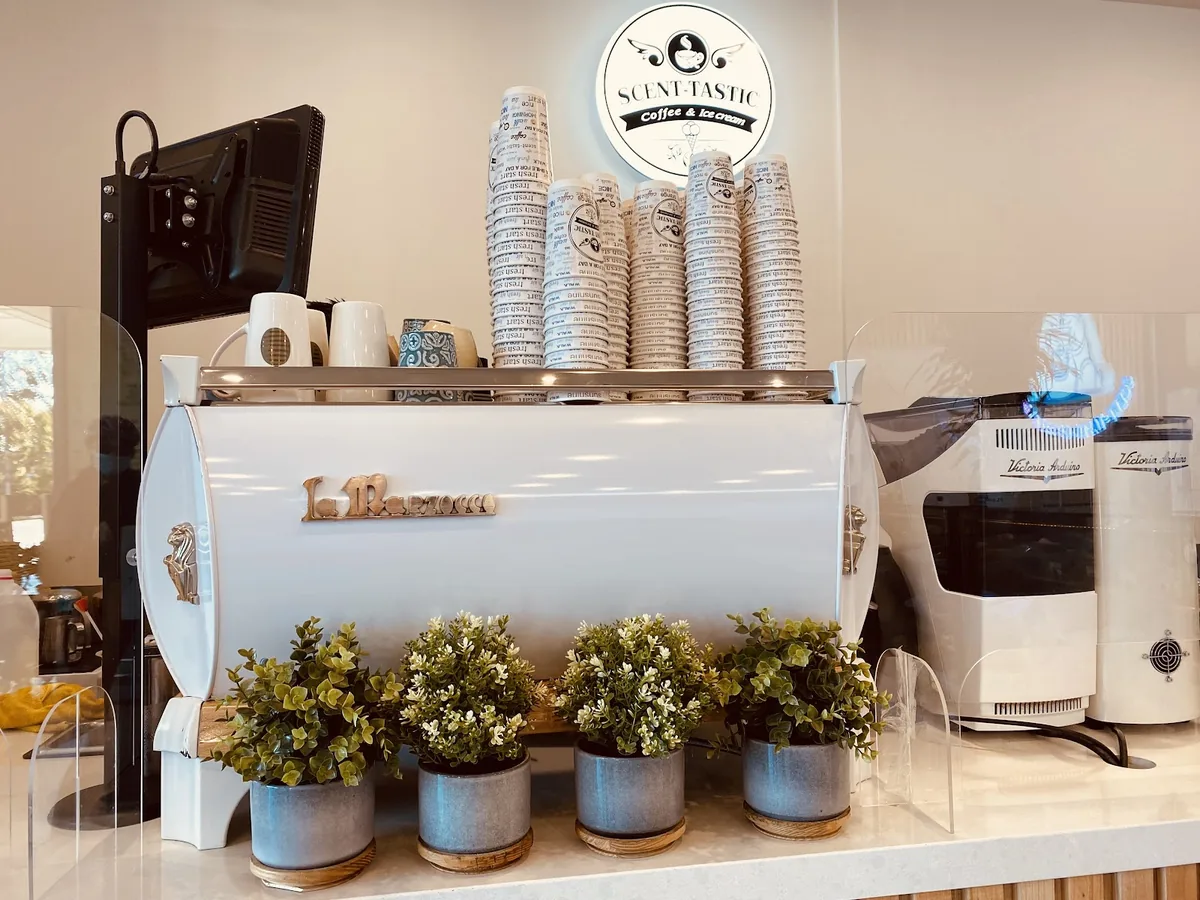 The white La Marzocco under the Scent-tastic sign, with a row of potted plants